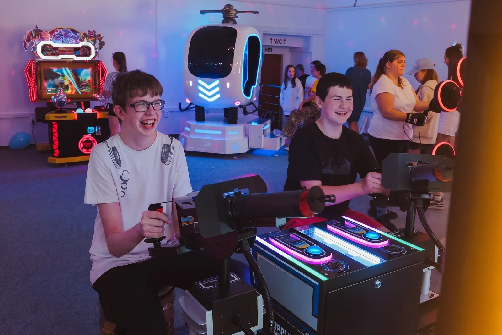 Dva rozesmátí kluci u herního automatu, v pozadí VR simulátor a další atrakce
